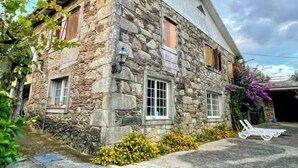 Ein Steinhaus mit einem geneigten Dach, zwei Fenster mit Fensterläden und ein kleiner Gartenbereich mit gelben Blumen.