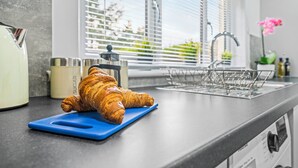 Un comptoir de cuisine avec un croissant posé sur une planche à découper bleue, un récipient à sucre, une machine à café et une fenêtre avec des stores.