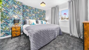 Une chambre à coucher comprenant un lit, une table de chevet, une lampe, une fenêtre avec des rideaux et un mur recouvert d’un papier peint à motifs tropicaux.