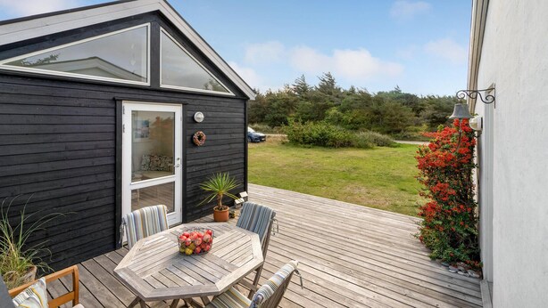 En lille, sort træhytte med en glasdør, en terrasse med bord og stole, og en have med røde blomster.