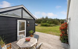 A small black wooden cabin with a glass door, a deck with a table and chairs, and a garden with red flowers. - 3 bedroom nice home in Ulfborg (Ulfborg)