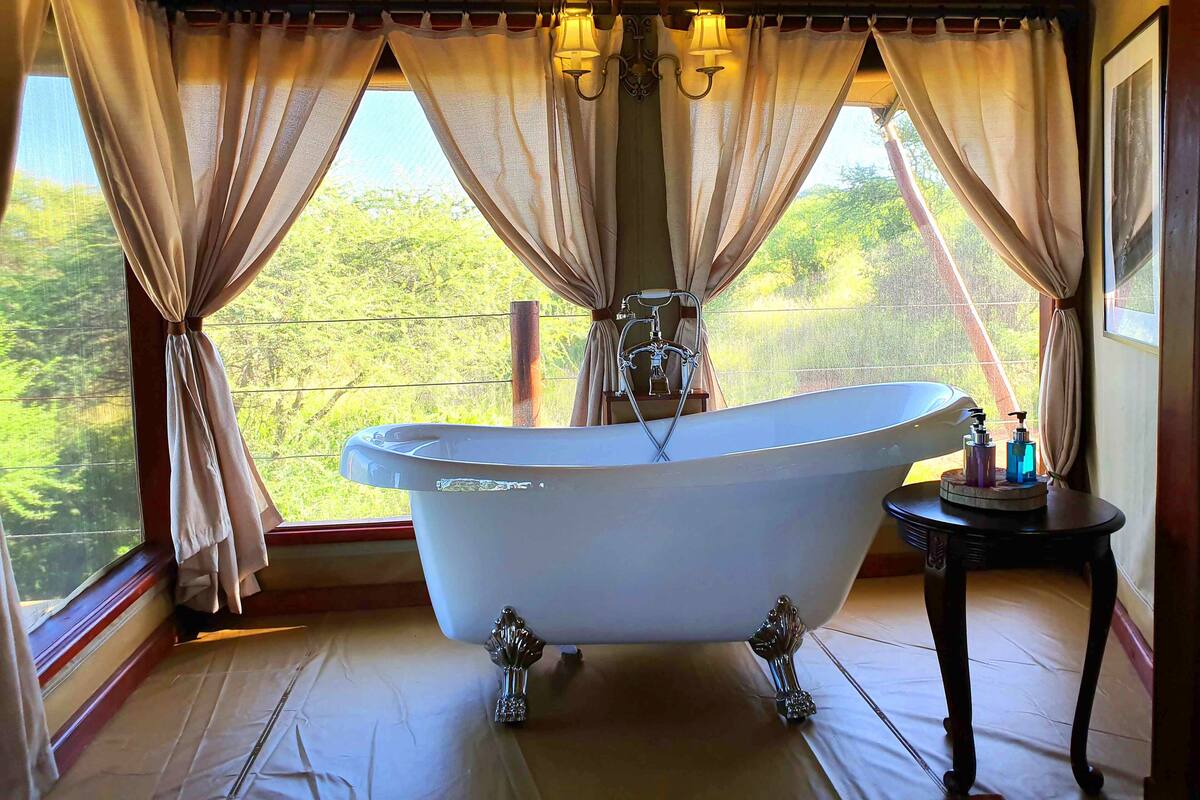 A bathroom with a freestanding bathtub, a small table with toiletries, and a view of greenery outside.