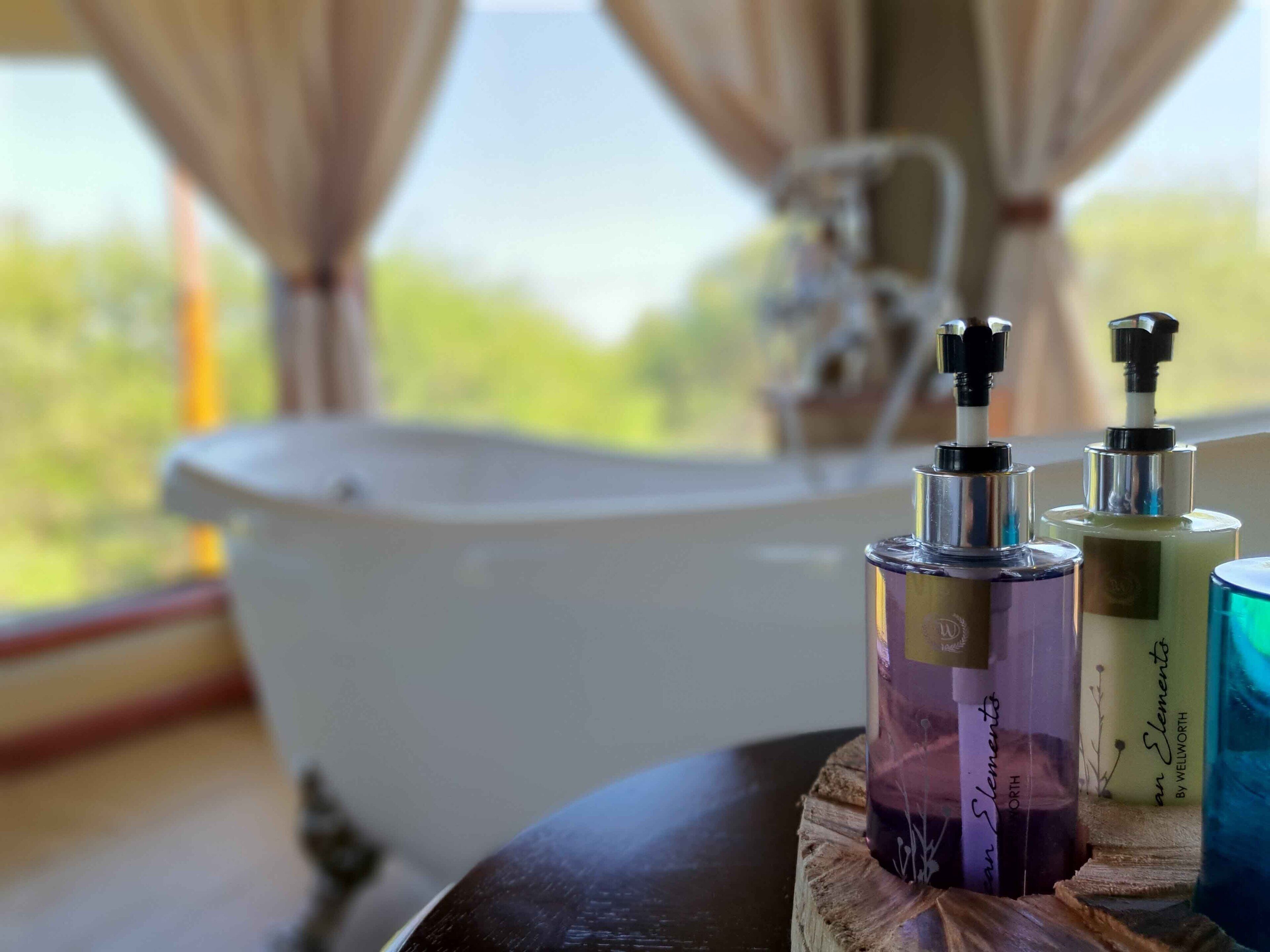 A bathroom with a bathtub, a wooden table with three bottles of toiletries, and a window with curtains.