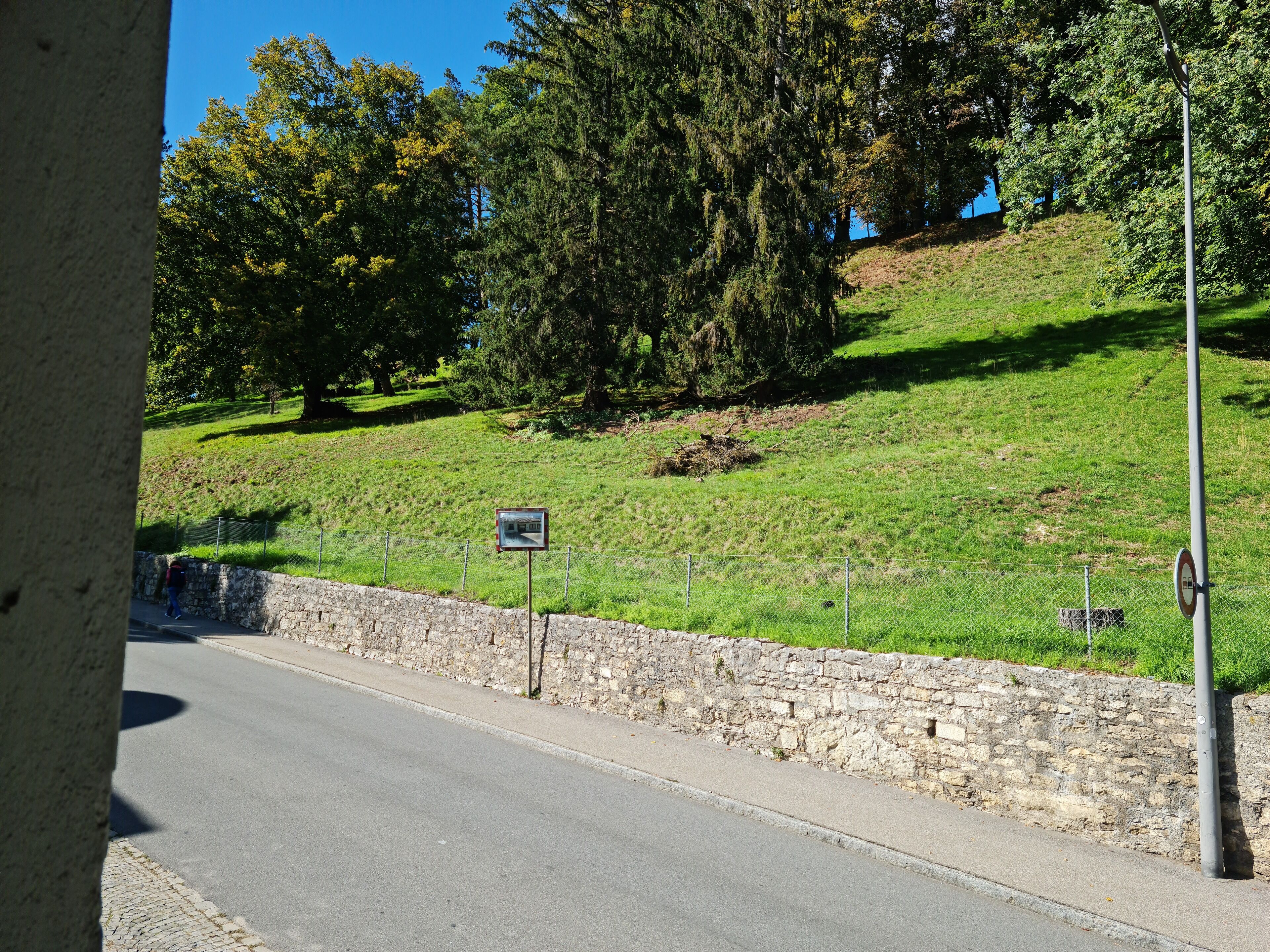 Un muro de piedra al lado de una carretera, con una valla metálica, un cartel y árboles al fondo.