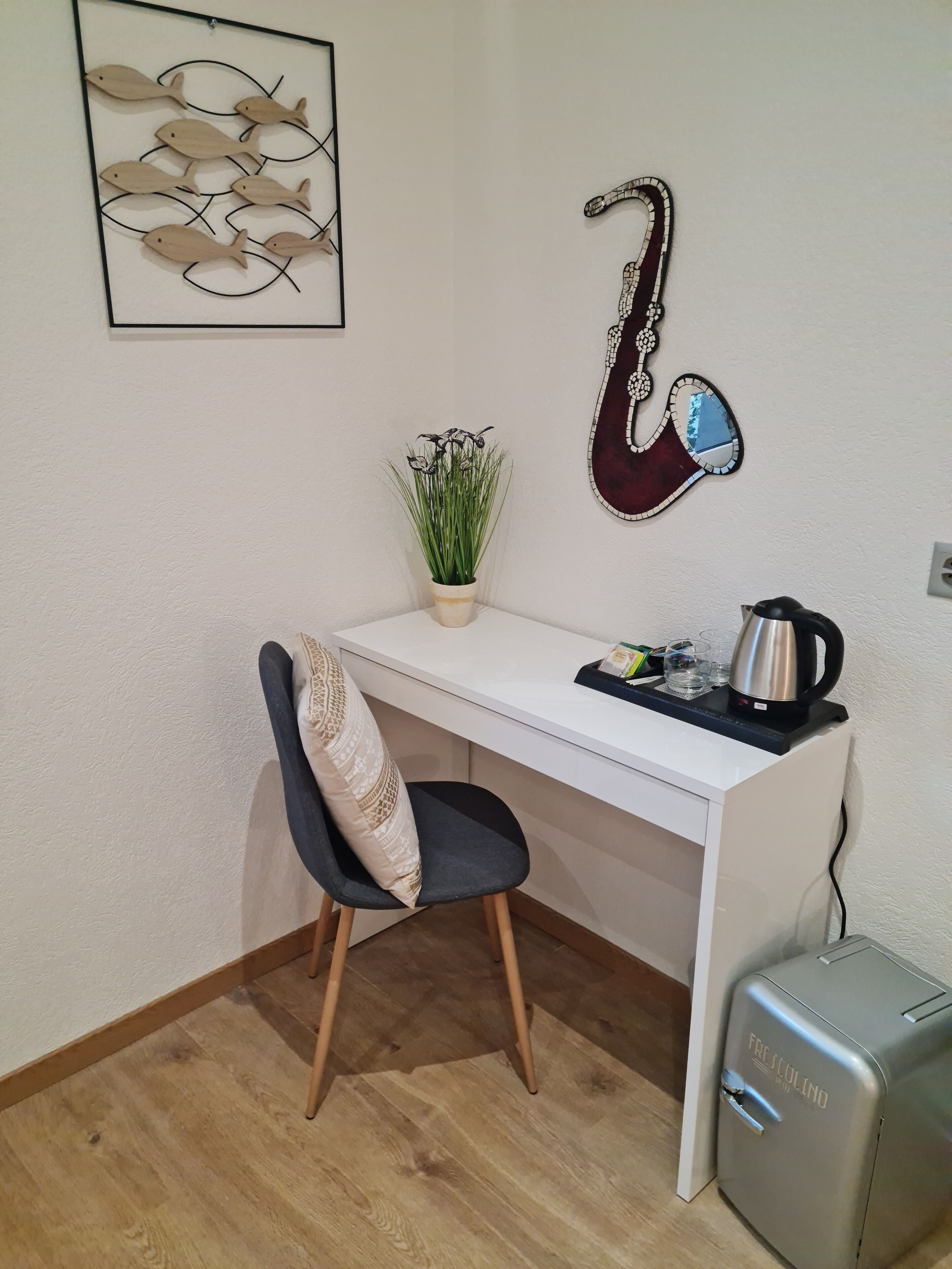 Quarto pequeno e bem organizado, com uma escrivaninha branca, uma cadeira, um vaso de planta, uma guitarra decorativa e um frigorífico compacto.