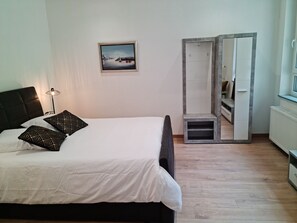A bedroom with a bed, a bedside lamp, a framed picture on the wall, and a wardrobe with a mirror. - LA FACTORY by LNA (Porrentruy)