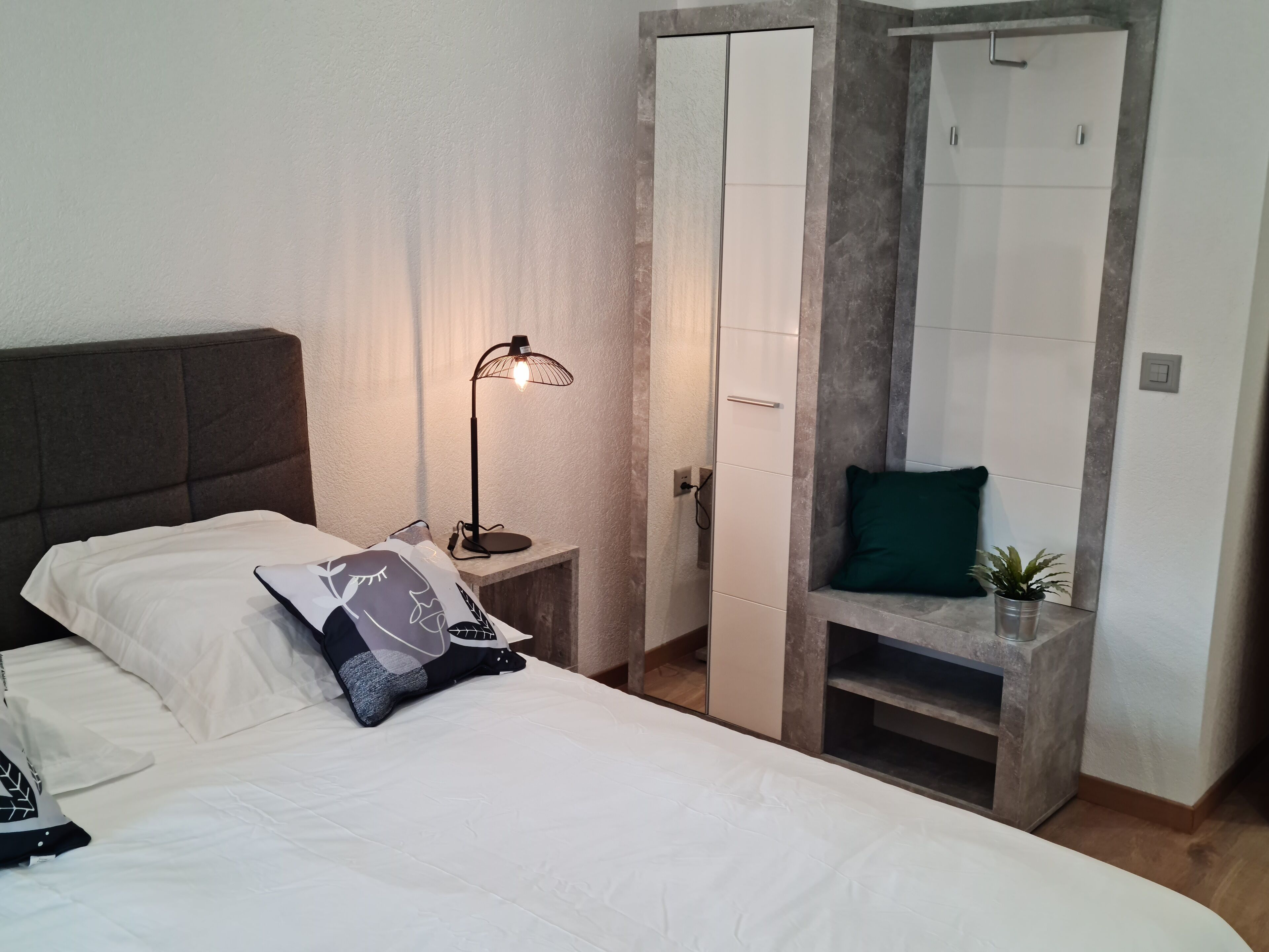 A neatly made bed with white linens and a single decorative pillow. A black floor lamp with a wire base is positioned on a small bedside table. A green pillow rests on a shelf against the wall, and a potted plant is placed on the floor beside the bed.