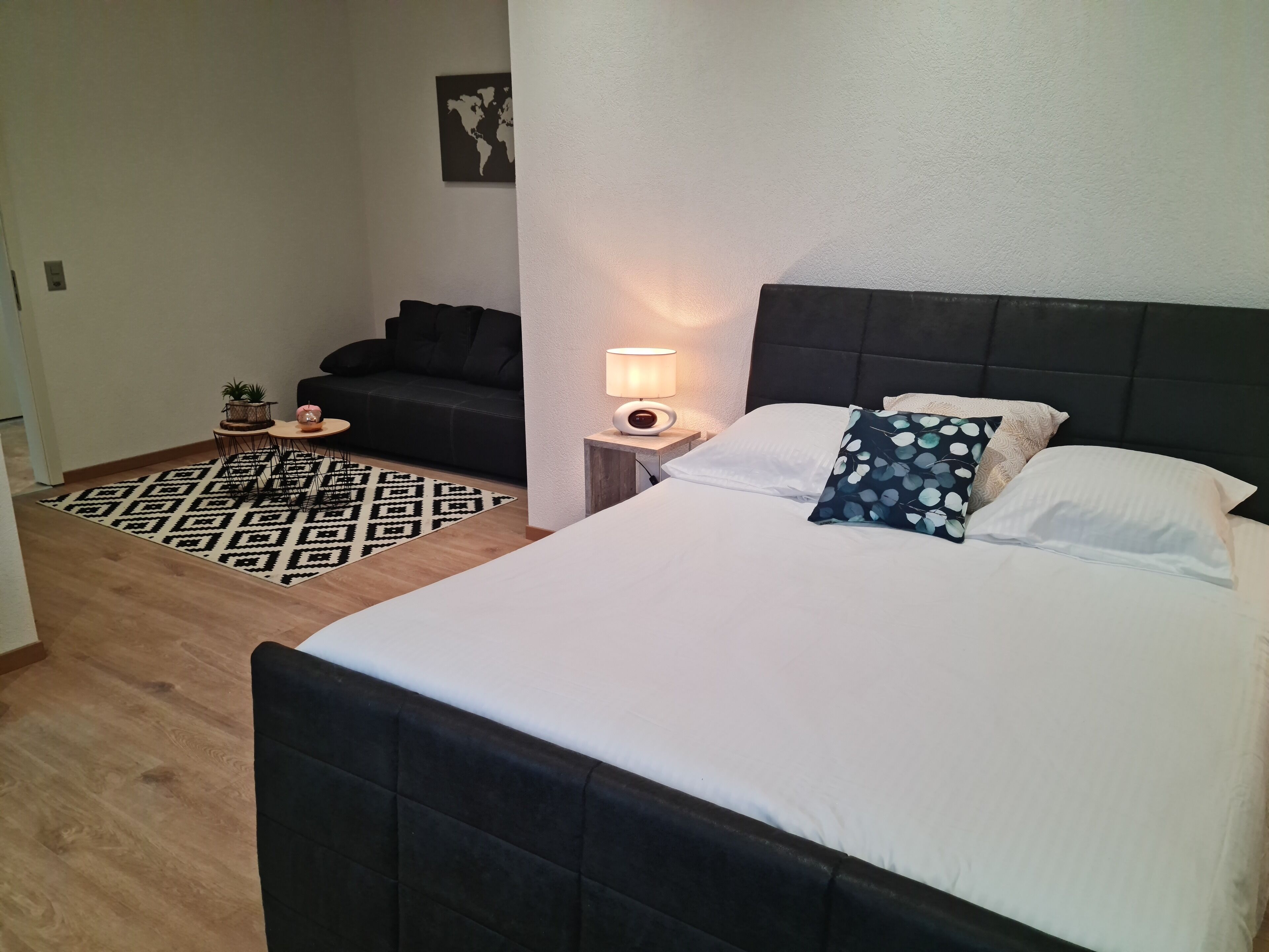 A hotel room with a bed, a nightstand with a lamp, a black sofa, a patterned rug, and a small table with a plant.