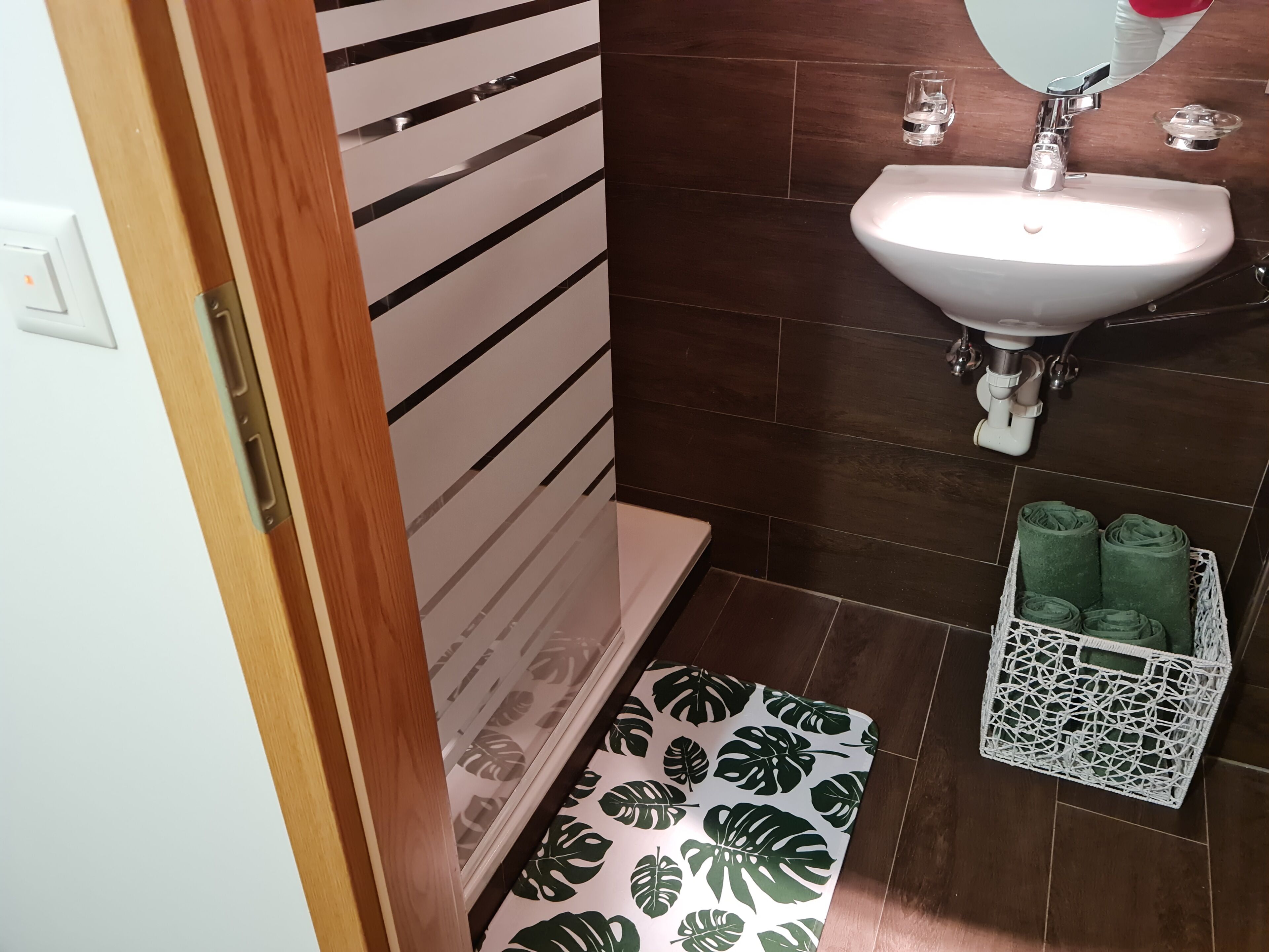 A bathroom with a white sink, dark brown tiles, a wooden door, and a white mat with green leaf patterns.