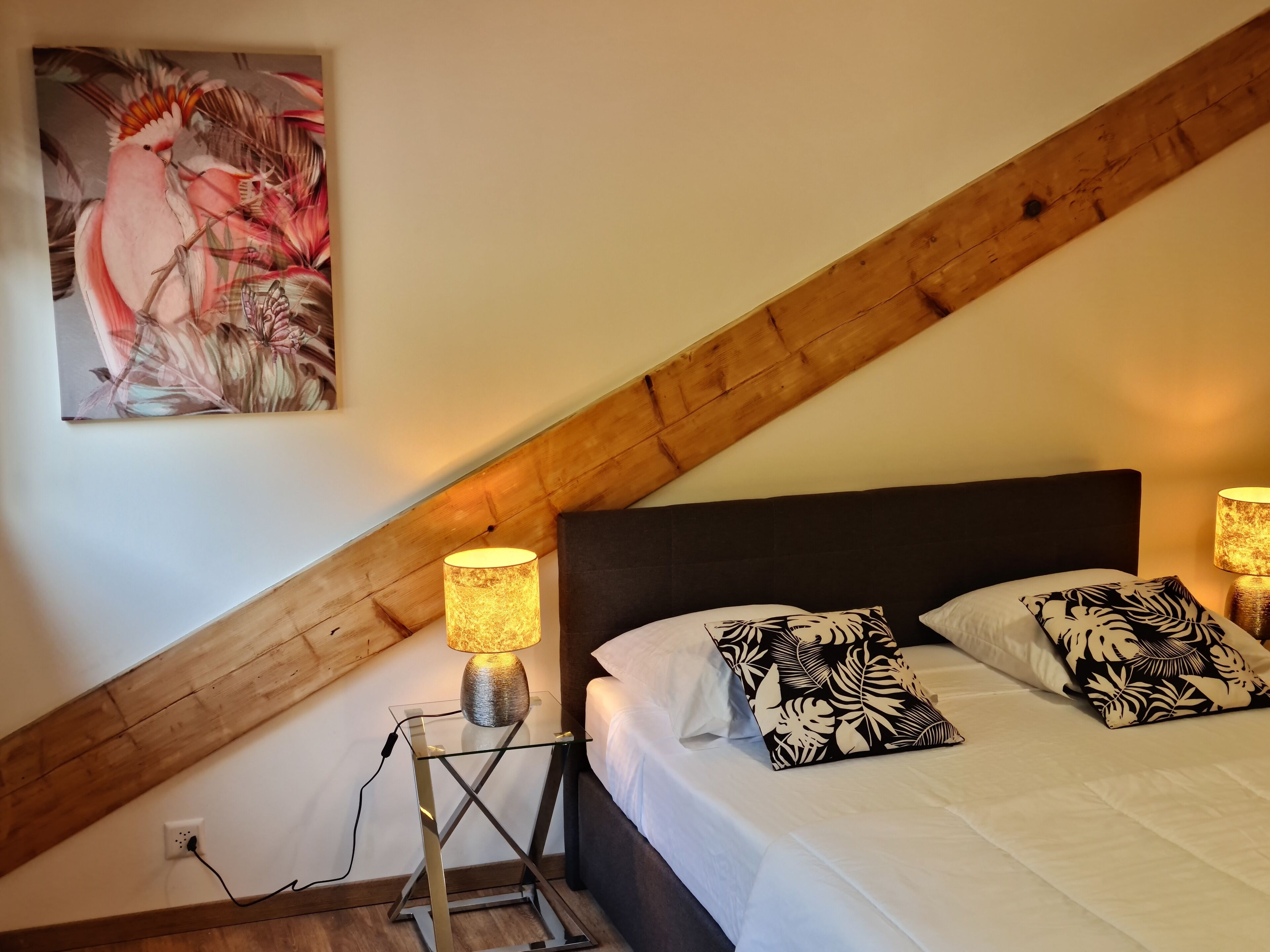 Un dormitorio con techo inclinado, una cama con almohadas estampadas, una mesita de noche con lámpara y un cuadro con motivos florales en la pared.