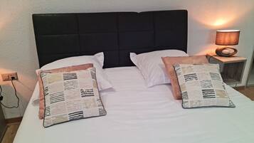 Una cama bien hecha con sábanas blancas y dos almohadones decorativos, un cabecero tapizado en negro, una lámpara de noche y un enchufe en la pared.
