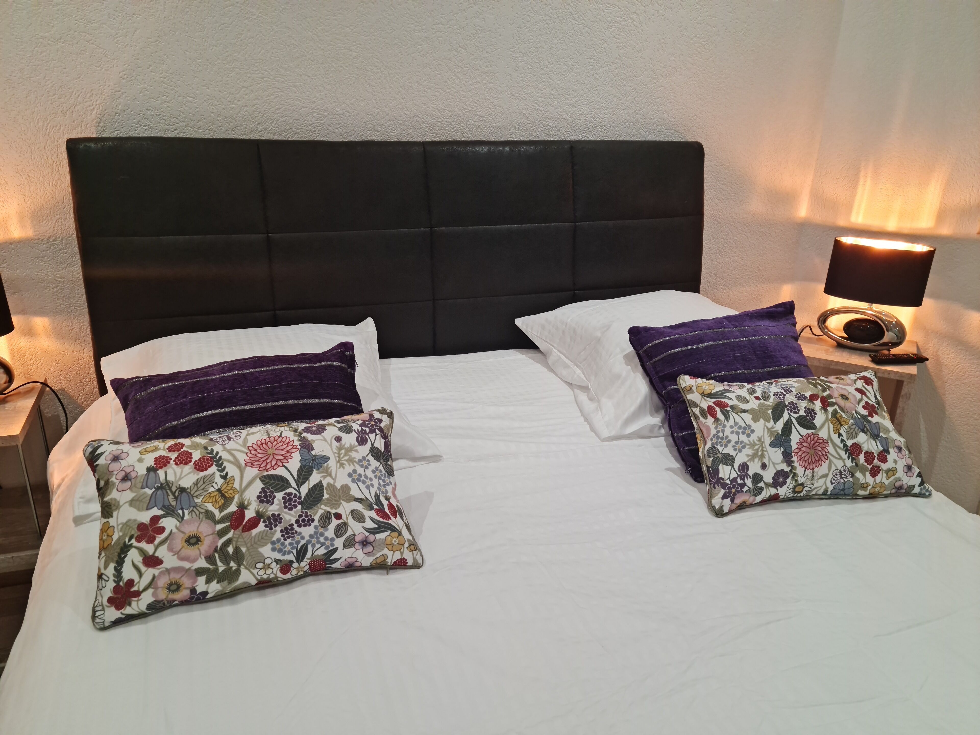 Una cama con cabecera negra, ropa de cama blanca y dos almohadas estampadas con motivos florales.