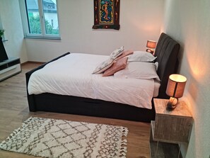 A neatly made bed with white linens and decorative pillows, a wooden headboard, a bedside lamp, and a patterned rug on a wooden floor. - LA FACTORY by LNA (Porrentruy)