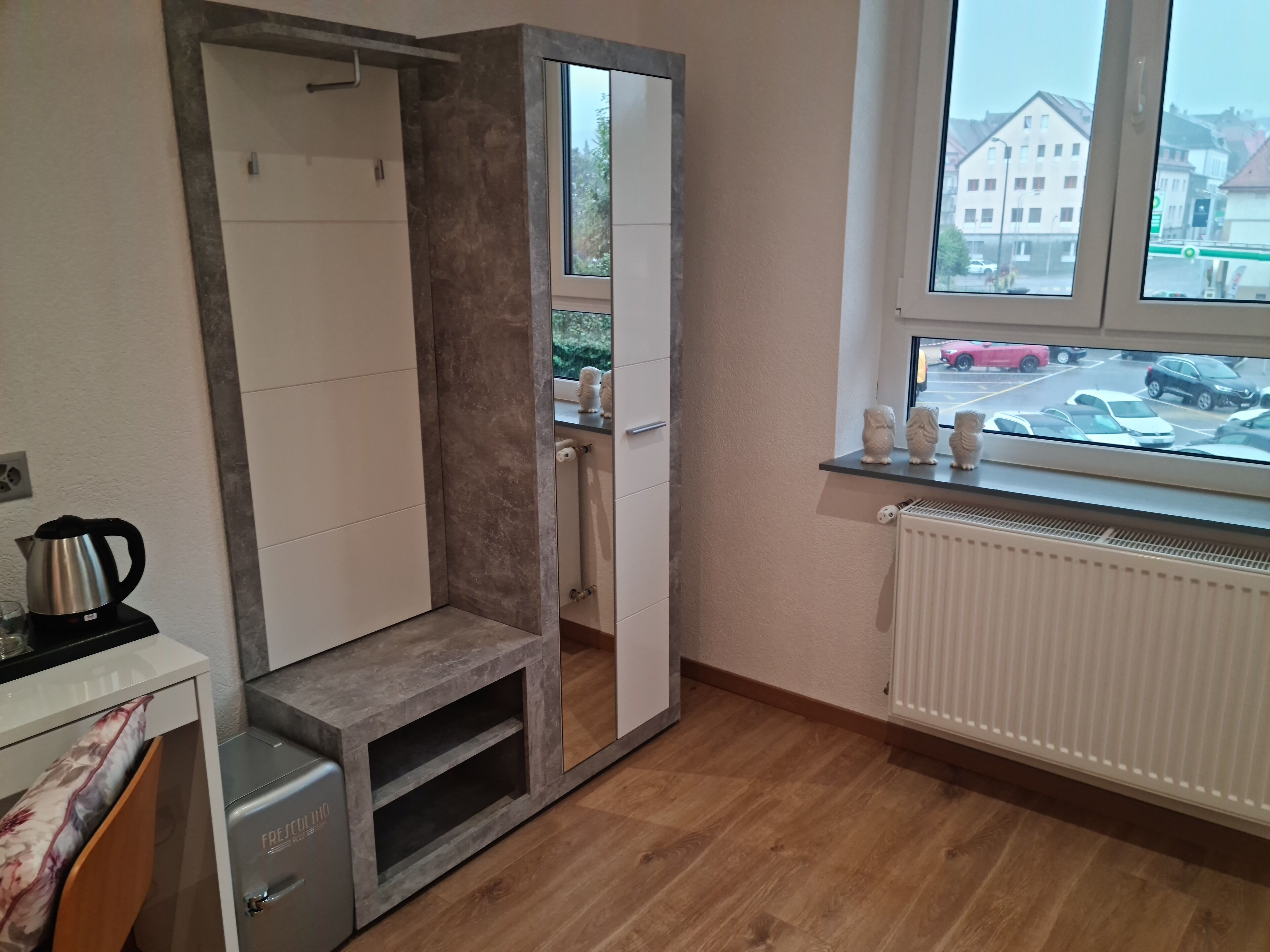 A compact room with a built-in wardrobe, a radiator, a small heater, a kettle, and a window with a view of parked cars and buildings.