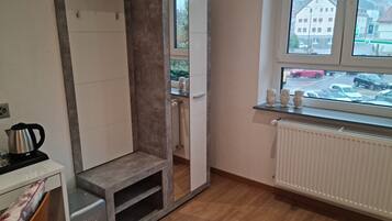 A compact room with a built-in wardrobe, a radiator, a small heater, a kettle, and a window with a view of parked cars and buildings.