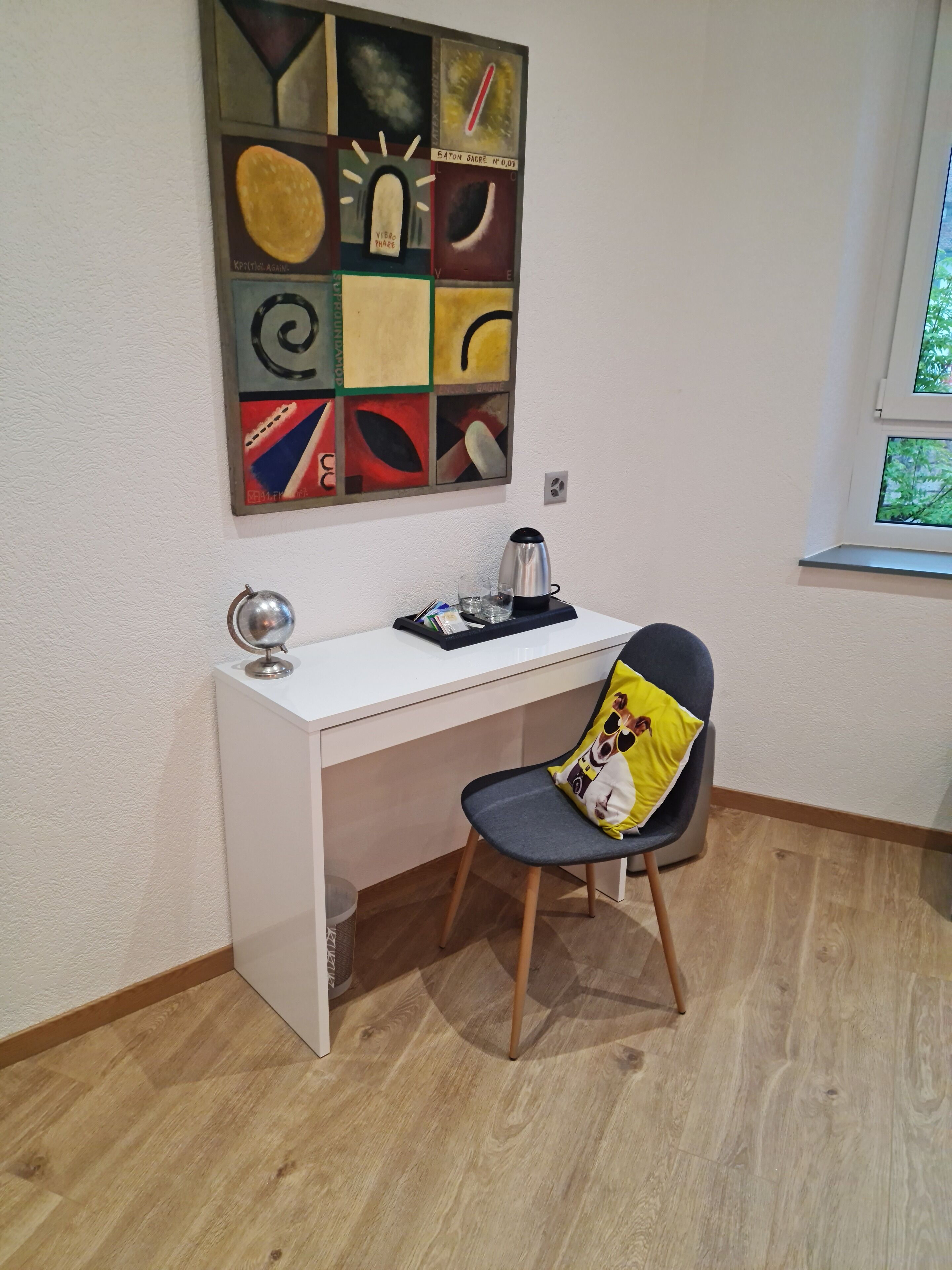 Una habitación pequeña y moderna con un escritorio blanco, una silla con un cojín de personaje de dibujos animados, un globo terráqueo, un hervidor y una ventana con vistas a árboles.