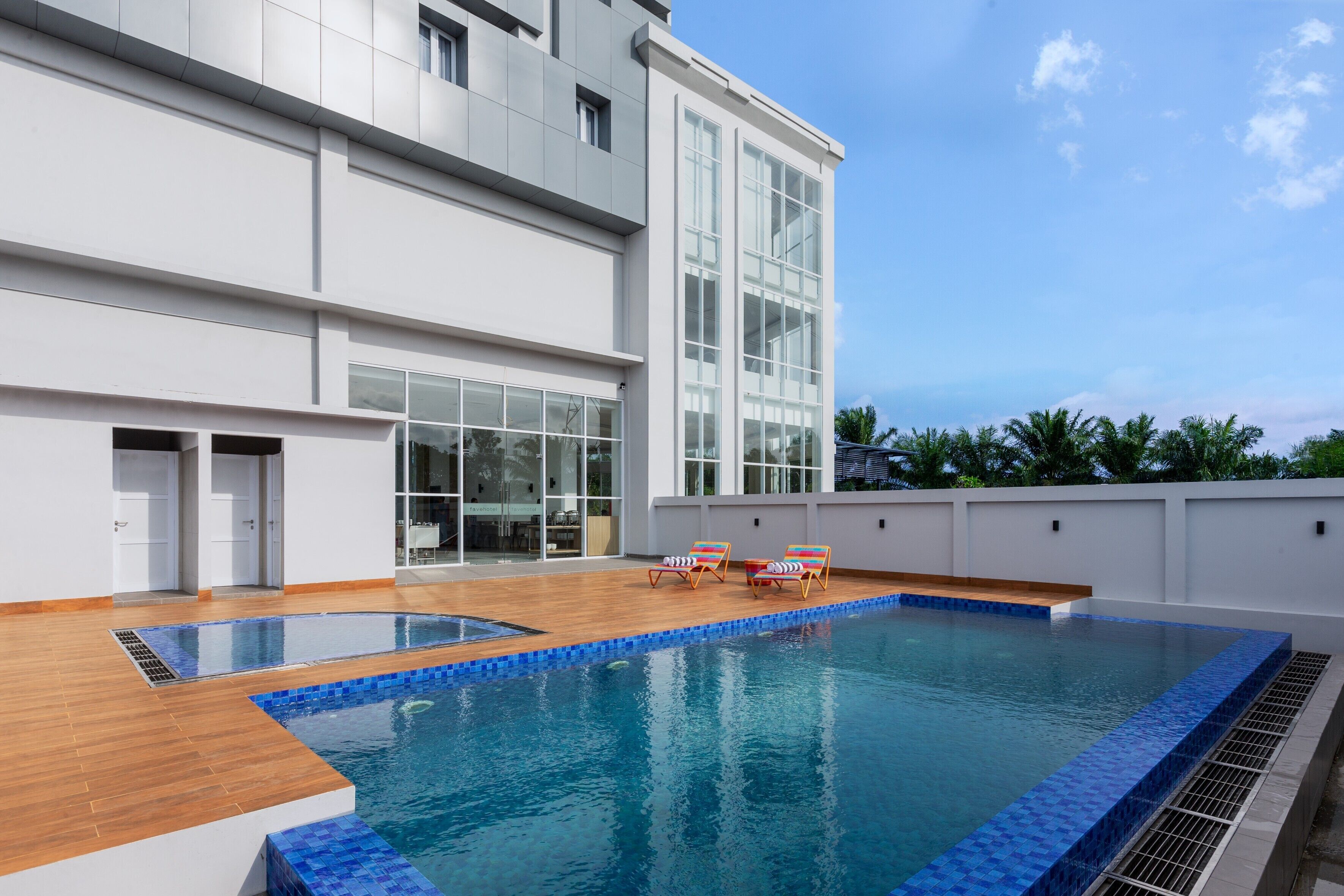 A rooftop swimming pool with wooden decking, two lounge chairs, and a modern building with large windows.