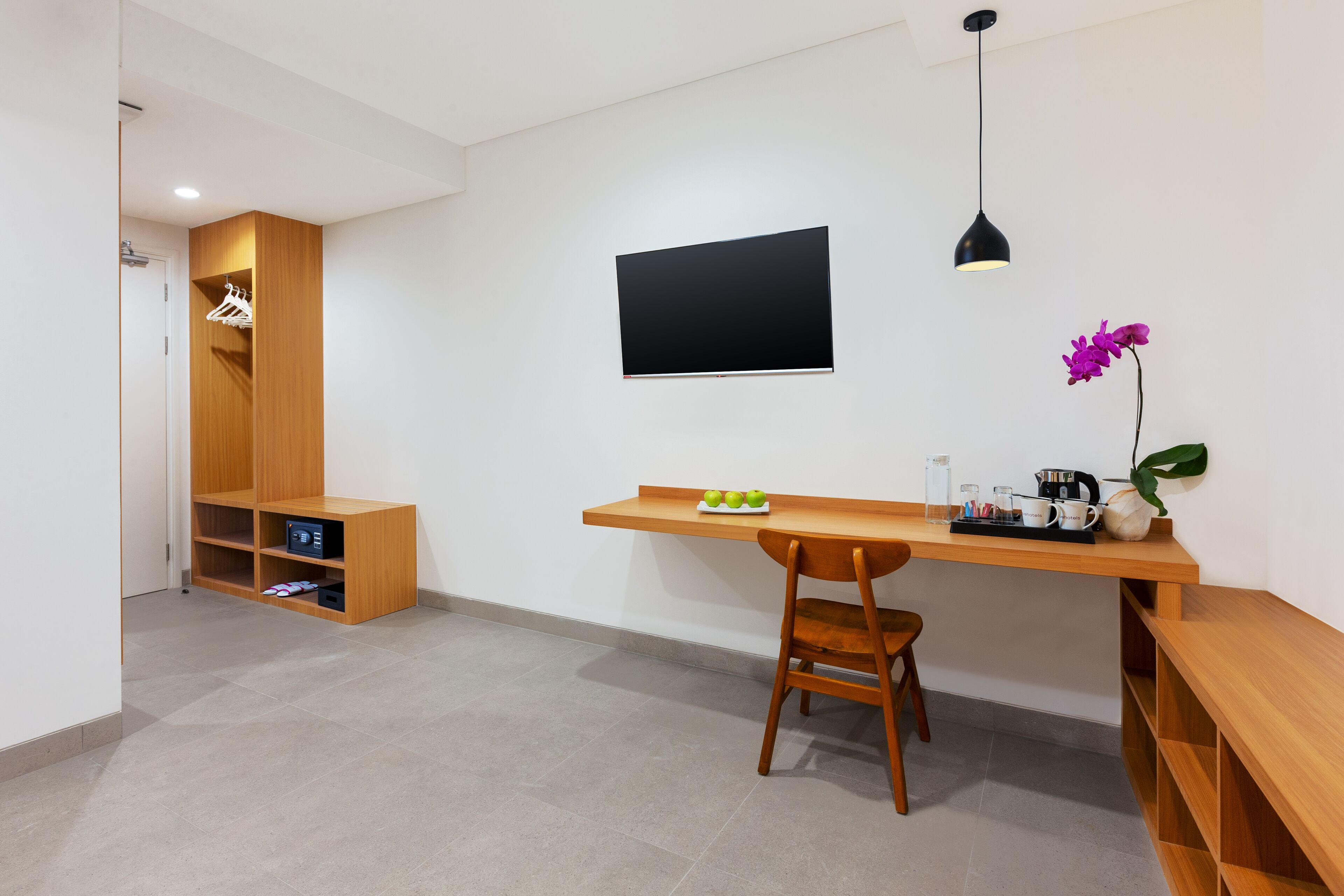 Una habitación moderna con un escritorio de madera, una silla, un televisor montado en la pared, una lámpara colgante y un pequeño estante con una planta.