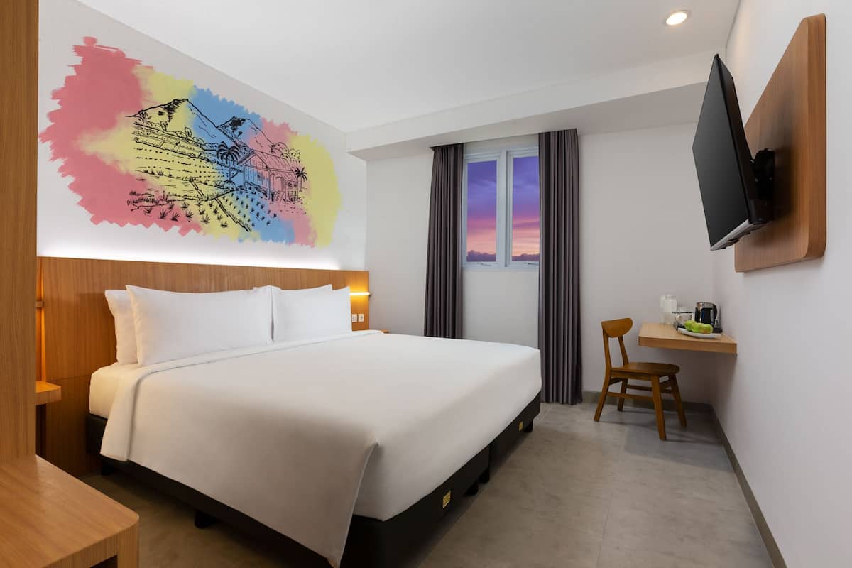 A hotel room with a bed, a desk with a chair, a TV mounted on the wall, and a window with curtains.