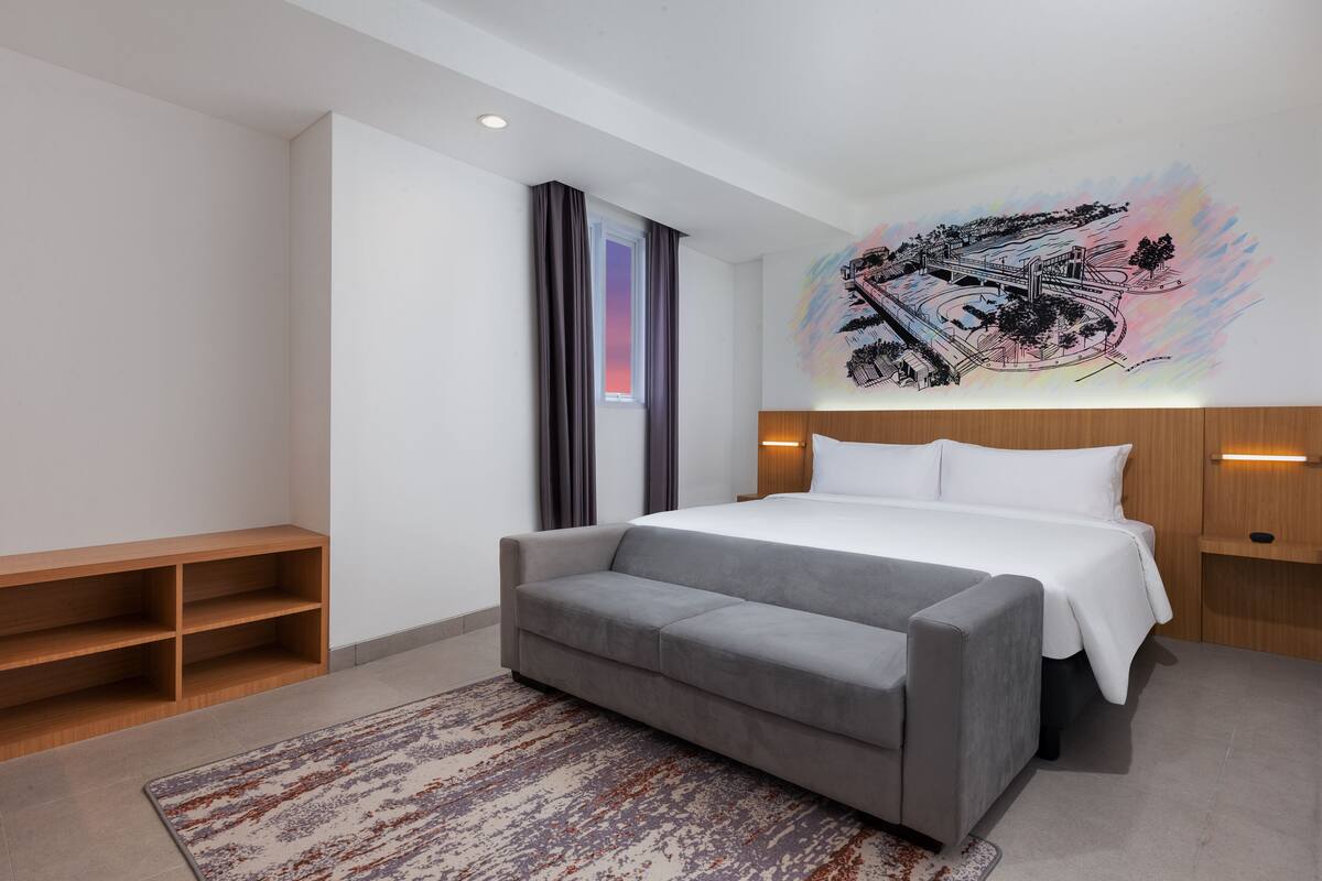 A modern hotel room with a large bed, a grey sofa, a wooden nightstand, and a colorful abstract painting on the wall.