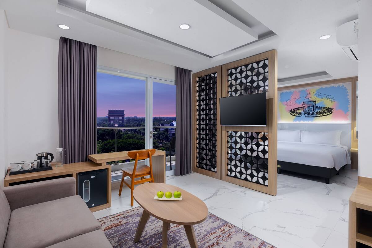 A modern hotel room with a large window, a bed, a desk with a chair, a small table with apples, and a TV on the wall.