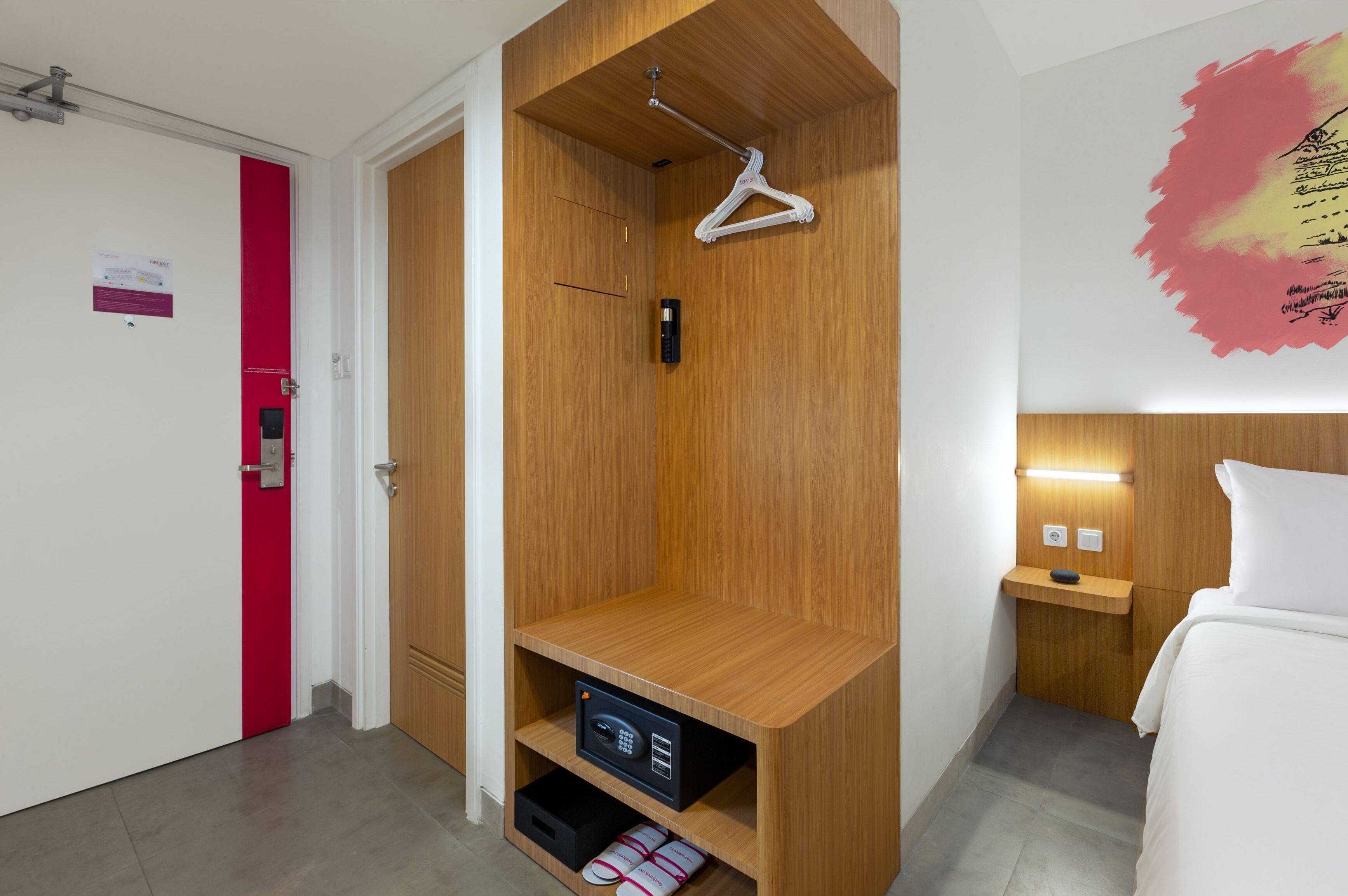 A hotel room with a wooden wardrobe, a hanging closet, a bedside table with a lamp, and a bed with white bedding.