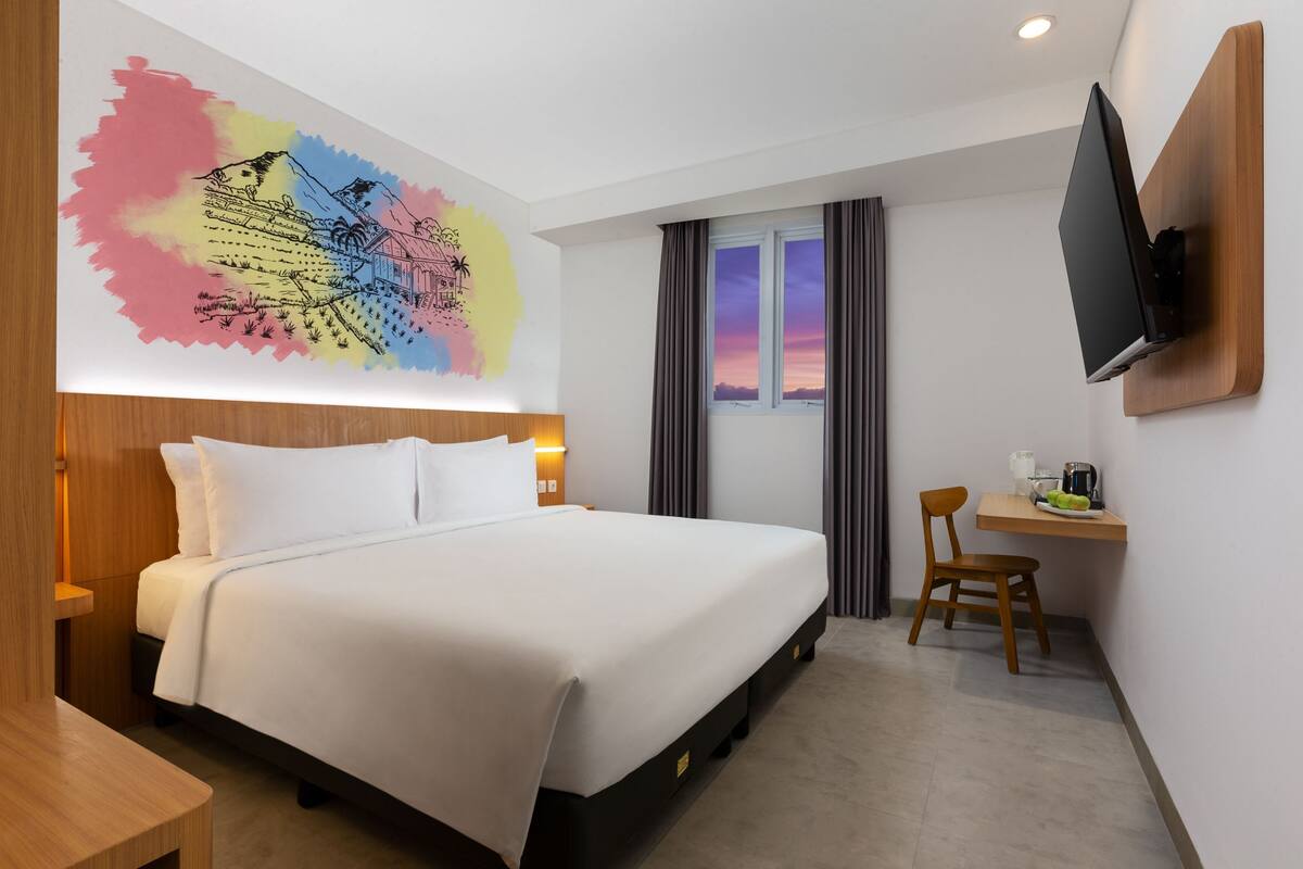 A hotel room with a bed, a desk with a chair, a TV mounted on the wall, and a window with curtains.