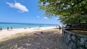 Una playa con agua azul cristalina, gente relajándose y barcos a lo lejos.