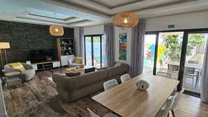 A modern hotel room with a wooden floor, a sofa, a dining table, a TV, and a balcony with a pool view. - Villa with pool 5 minutes from the beach! (Trou-aux-Biches)