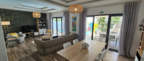 A modern hotel room with a wooden floor, a sofa, a dining table, a TV, and a balcony with a pool view.