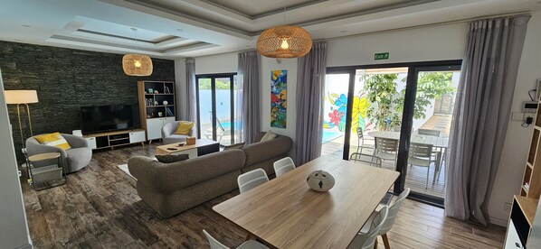 Una habitación de hotel moderna con piso de madera, un sofá, una mesa de comedor, un televisor y un balcón con vista a la piscina.