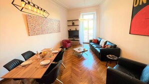 A modern living room with a wooden dining table, black leather sofas, and a large abstract painting on the wall. - MASSENA AP4312 By Riviera Holiday Homes (Niza)