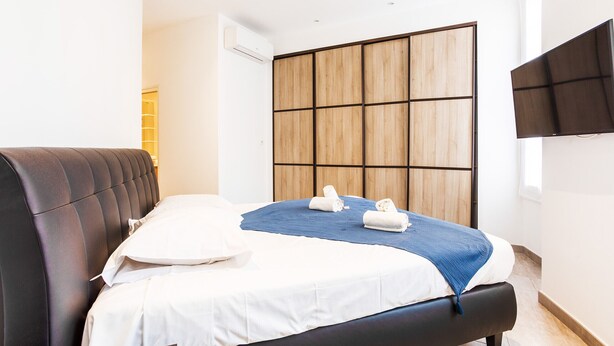 A modern hotel room with a large bed, a wooden panel wall, and a flat-screen TV. - MASSENA AP4312 By Riviera Holiday Homes (Niza)