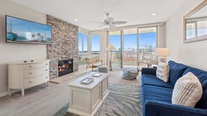A spacious living room with a stone fireplace, a large TV, a blue sofa, and a view of the ocean through large windows. - G38 Harbor Cottage at North Coast Village - Beautiful Ocean Views *Super Close to Beach* (Oceanside)