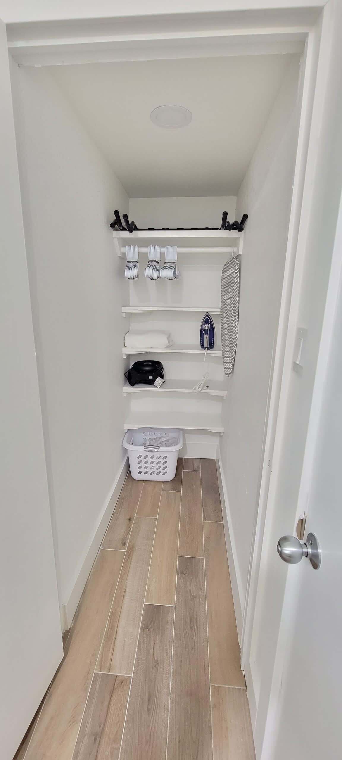A narrow hallway with white shelves containing laundry supplies, a laundry basket, and an ironing board.