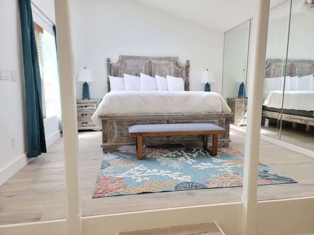 A bedroom with a large bed, a bench, a patterned rug, and a mirror.