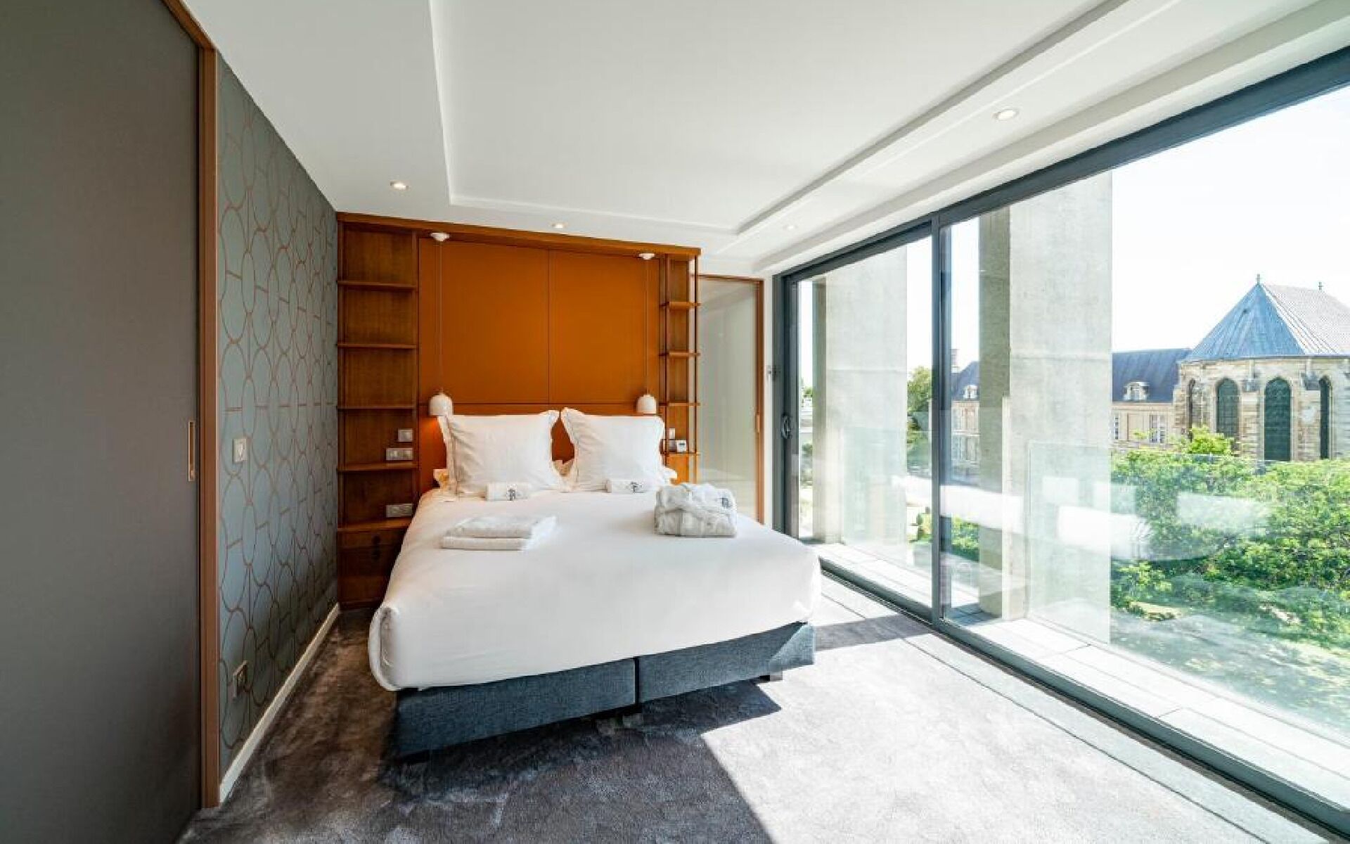 A modern bedroom with a large bed, built-in shelving, and a view of a building through large windows.