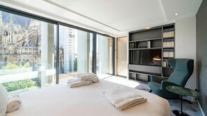 Une chambre d’hôtel moderne dotée d’un grand lit, d’un coin salon avec un fauteuil, d’une bibliothèque et offrant une vue sur un bâtiment historique grâce à de grandes fenêtres.