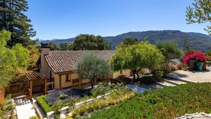 Una casa con techo de tejas, rodeada de un jardín con diversas plantas y vistas a una cadena montañosa.