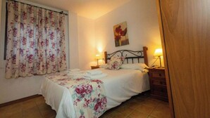 Un dormitorio con una cama, dos mesitas de noche con lámparas, un ventanal con cortinas florales y un cuadro en la pared.