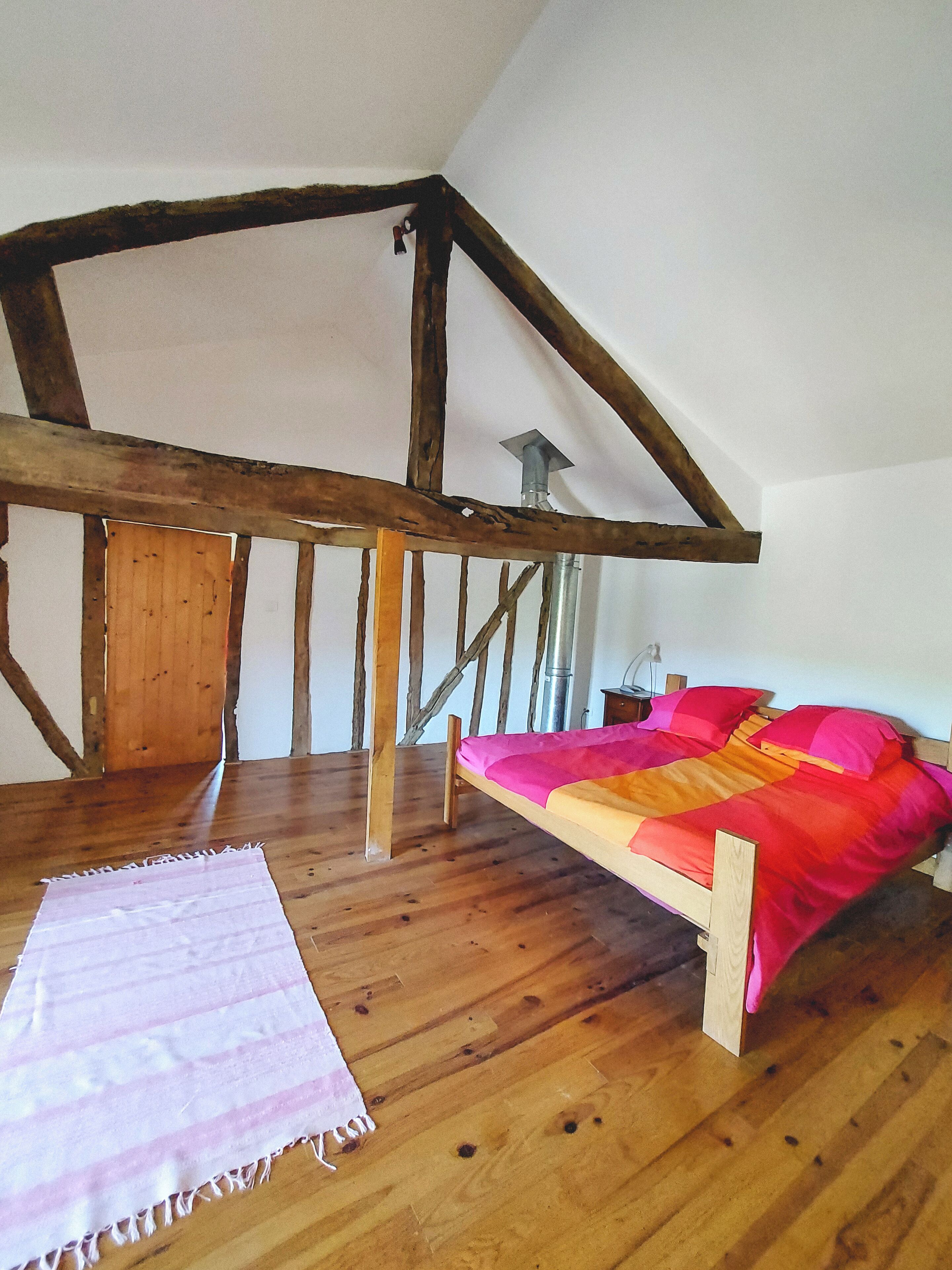 A bedroom with a sloped ceiling, wooden beams, a bed with a colorful blanket, and a wooden floor.