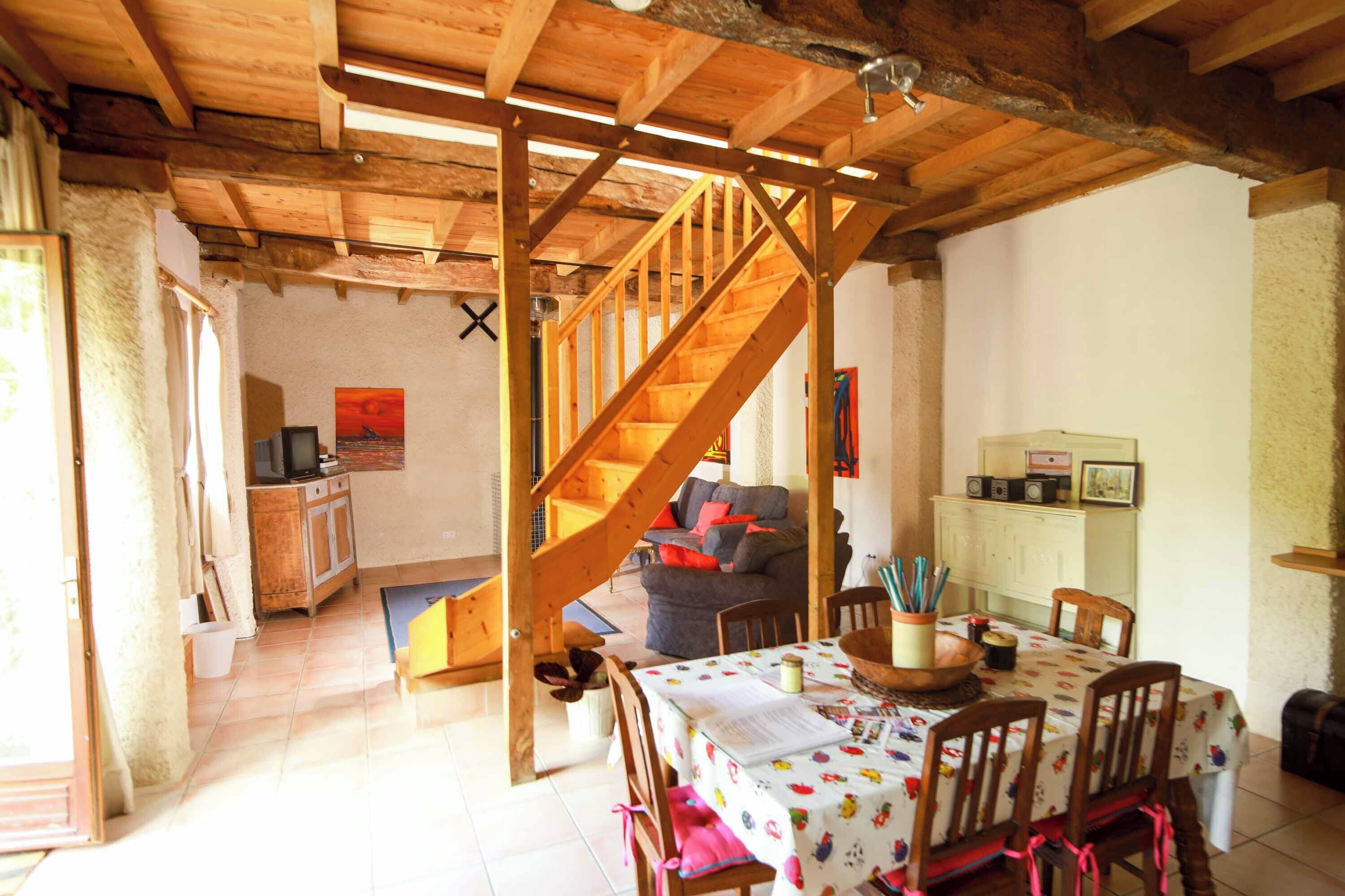 A rustic interior with a wooden staircase, a dining area with a table and chairs, and a living room with a sofa and television.