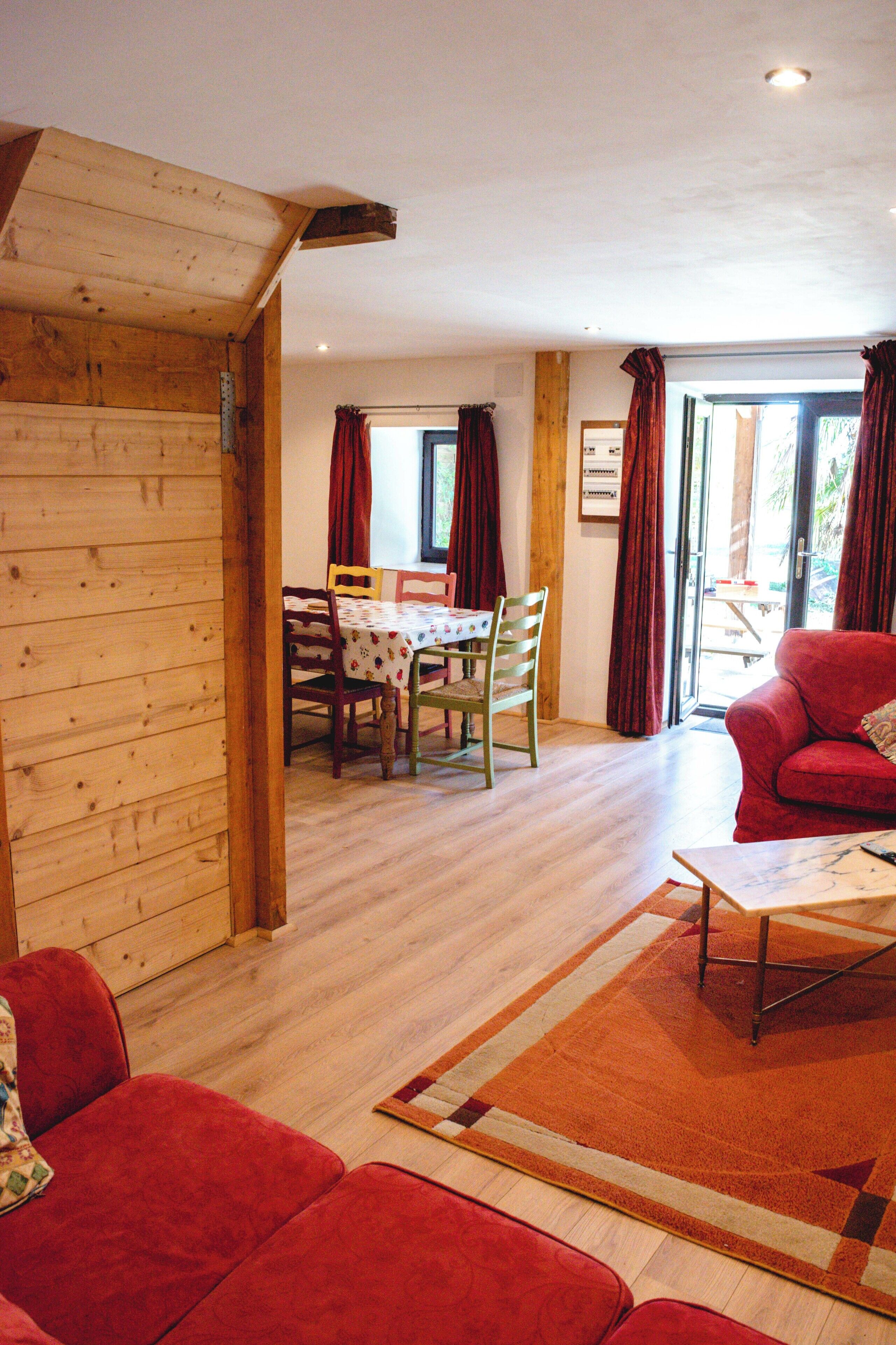 Een gezellige woonkamer met een houten wand, een rode bank, een eethoek met tafel en stoelen, en een groot raam met gordijnen.