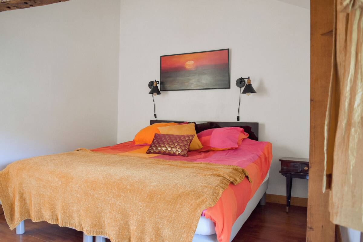Quarto com cama, almofadas, manta, pintura na parede e duas luminárias de cabeceira.