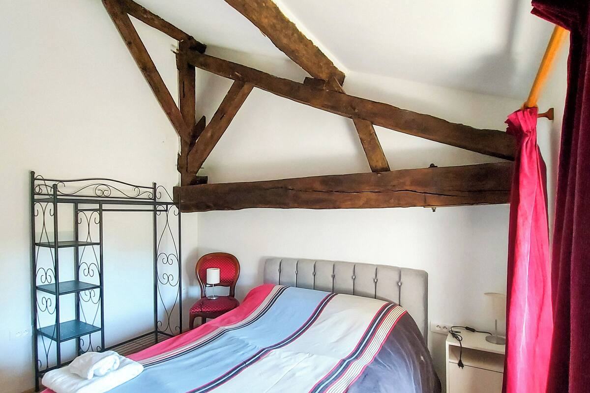Quarto com cama, mesa de cabeceira, cadeira, guarda-roupa e teto com vigas de madeira aparente.