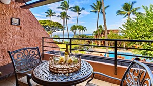 A round table with a cake and champagne, two chairs, a stone wall, and a view of a pool and palm trees. - Tropical vibes, sleek style, a peek at the Ocean (Lahaina)