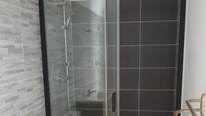Moderni kylpyhuone, jossa on lasiseinällä varustettu suihku, valkoinen wc-istuin ja puinen allaskaappi.
