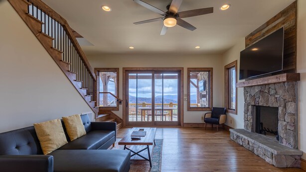 A spacious living room with a black leather sofa, a wooden coffee table, and a stone fireplace. A ceiling fan and a flat-screen TV are mounted on the wall.