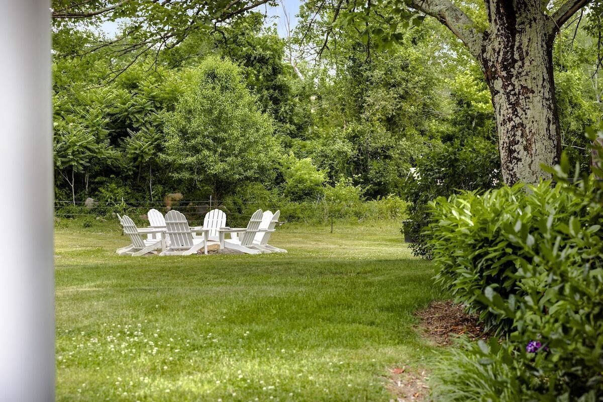 Un espacio al aire libre cubierto de césped, con tres sillas Adirondack blancas y una mesa, rodeado de densa vegetación verde.