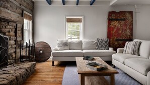 A living room with a stone fireplace, a white sofa, a wooden coffee table, and a large window with a view of greenery. - Walk to Old Rag | Hot Tub | Dog-Friendly Farmhouse (Sperryville)